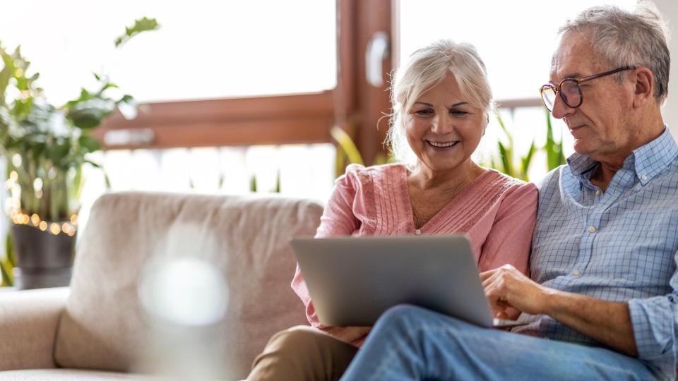 Ein älteres, lächelndes Paar sitzt auf dem Sofa und schaut auf einen Laptop