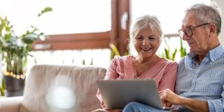 Ein älteres, lächelndes Paar sitzt auf dem Sofa und schaut auf einen Laptop